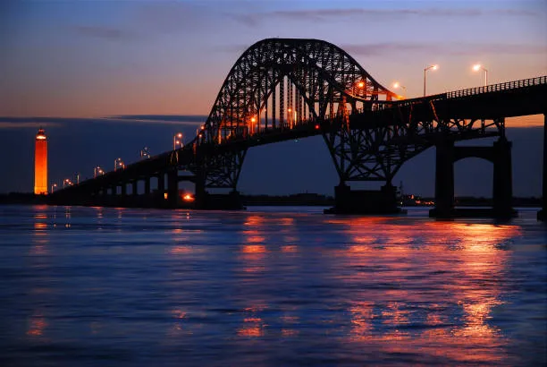 Steel arch bridge at dusk over calm water, with an orange lighthouse on the left and reflections of bridge lights on the surface, creating a warm scene.