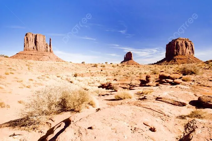 Desert panorama with three large sandstone buttes rising from sandy foreground under a clear blue sky and sparse desert shrubs alternating with rocky ground.